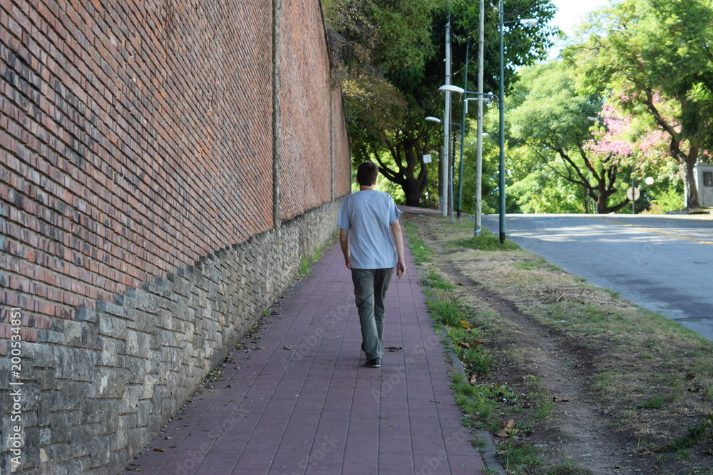 the man walks along the road along the brick wall which rises up the hill