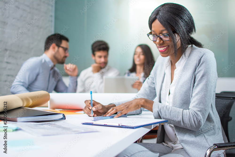 Smiling young business woman taking notes to notebook while sitting in office with colleagues in the background. Smiling young business woman taking notes to notebook while sitting in office with colleagues in the background.