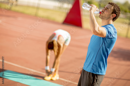 Fototapeta Naklejka Na Ścianę i Meble -  Sporty man drinking water while young woman doing exercise in the background