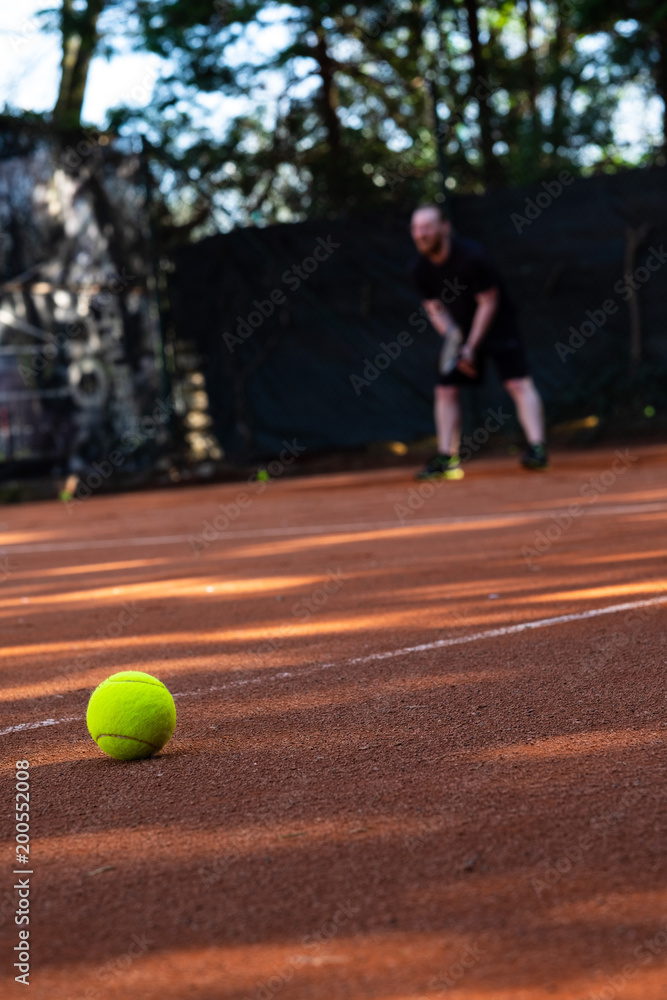 Closeup of tennis ball on a clay field with a blurred person playing in the background