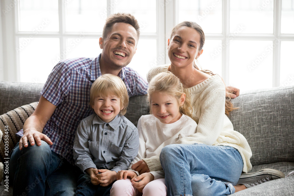 Portrait of happy multinational family with adopted kids laughing bonding together, smiling young couple embracing boy son and daughter girl sitting on couch, children and parents looking at camera Portrait of happy multinational family with adopted kids laughing bonding together, smiling young couple embracing boy son and daughter girl sitting on couch, children and parents looking at camera