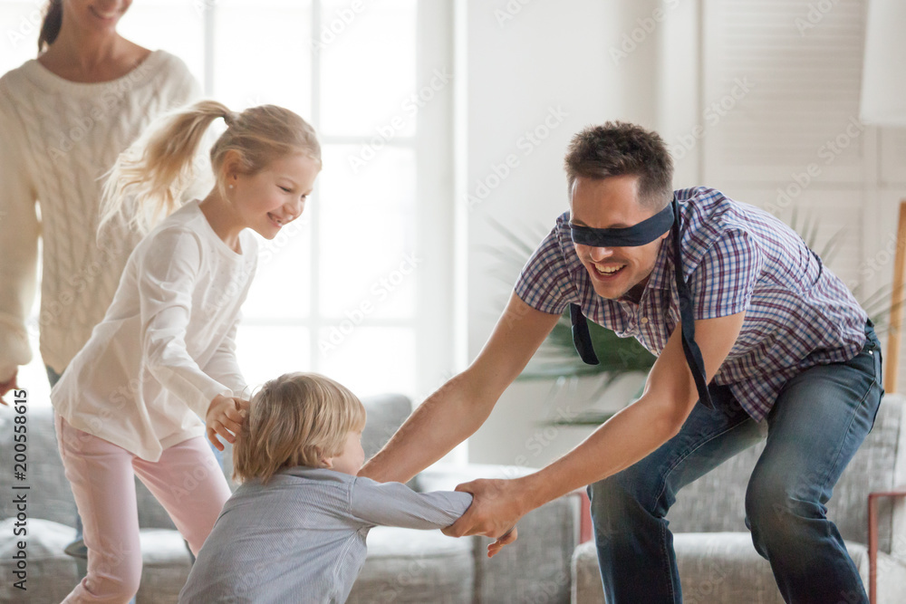 Happy young family of four playing hide and seek game at home, blindfolded father trying to catch children escaping, parents and kids having fun laughing spending free time together in living room Happy young family of four playing hide and seek game at home, blindfolded father trying to catch children escaping, parents and kids having fun laughing spending free time together in living room