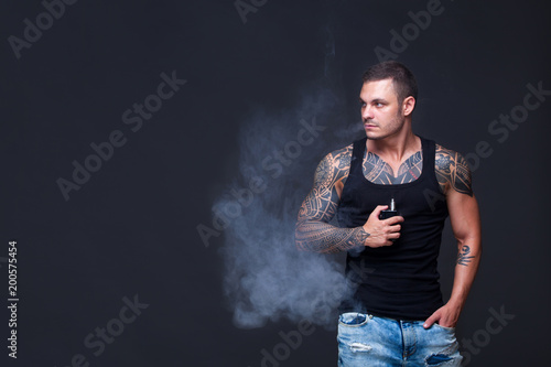 Vaper. The man with a muscular torso with tattoos smoke an electronic cigarette on the dark background photo