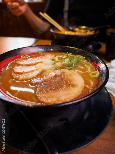 Close-up detail of a bowl of hot miso ramen topped with roasted chashu pork with customers eating in the background. Vertical orientation. Osaka, Japan. Travel and cuisine concept.