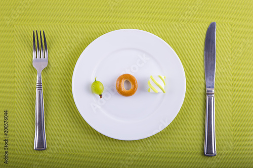 Minimalistic food composition with different types of products on white plate, knife and fork on green background