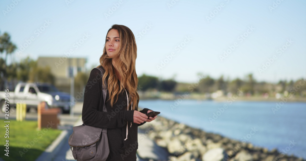 Young millennial girl messaging on cell phone by the bay smiling and laughing