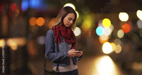 Trendy millennial girl messaging on smartphone on city street at night