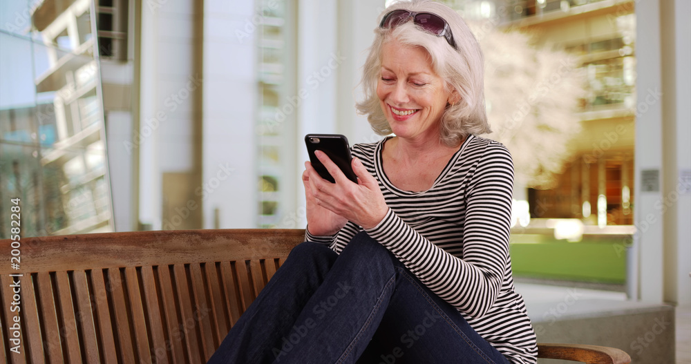 Smiling Caucasian female senior using smartphone while sitting outside on public bench Smiling Caucasian female senior using smartphone while sitting outside on public bench