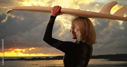 Photography Mature white female surfer holding surfboard over head watching sunset at beach