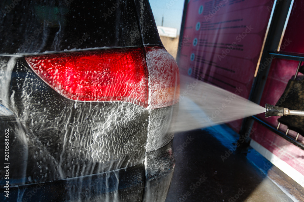 Naklejka premium Jet of wash with soap water high pressure is sent to the rear lights of the machine. Driver, girl wahsing her car in the stall of a car wash, using a high pressure water jet.
