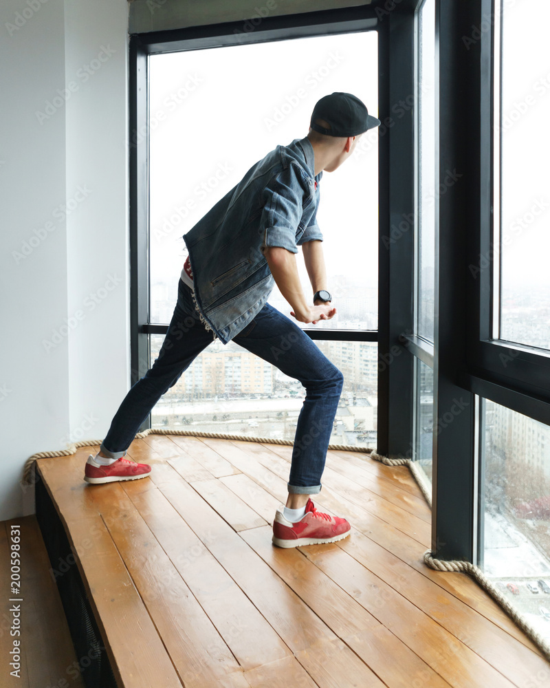 Teenage boy in denim jacket and baseball cap, jeans and sneakers dances by window in dance studio. Hip-hop movement. Dynamics of modern dance Teenage boy in denim jacket and baseball cap, jeans and sneakers dances by window in dance studio. Hip-hop movement. Dynamics of modern dance