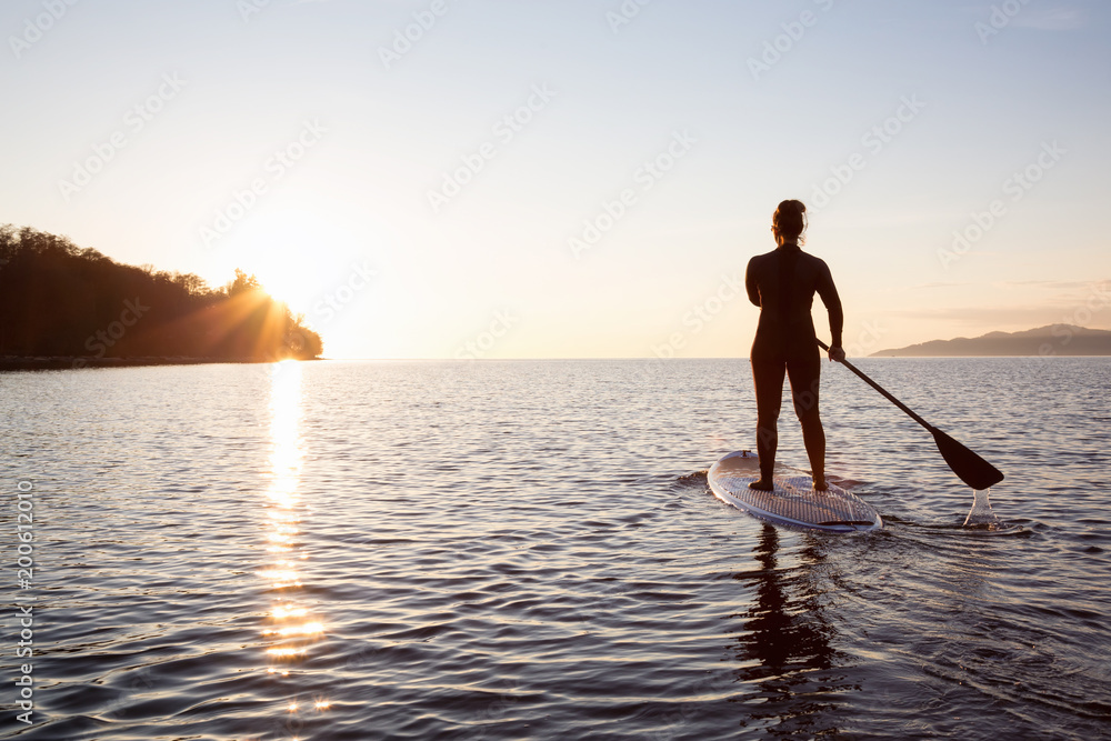 Adventurous girl on a paddle board is paddeling during a bright and vibrant sunset. Taken near Spanish Banks, Vancouver, British Columbia, Canada.
