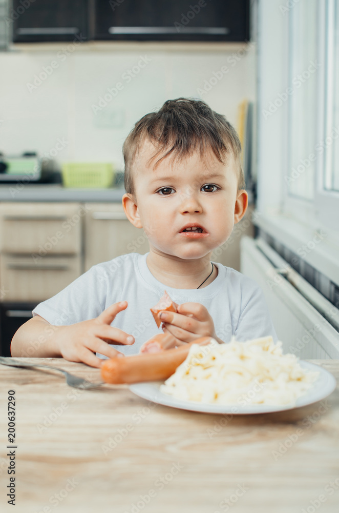The child is sitting in the kitchen and greedily eats sausage The child is sitting in the kitchen and greedily eats sausage