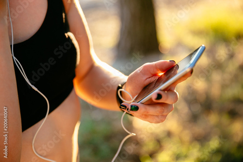 Close-up photo of sportive woman in black tank top using a mobile phone while listening to music in earphones after workout in the park.