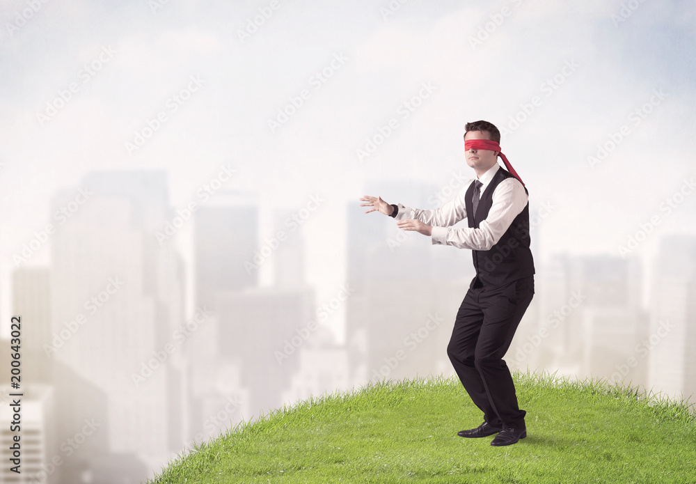Young blindfolded businessman steps on a a patch of grass with a city in the background Young blindfolded businessman steps on a a patch of grass with a city in the background