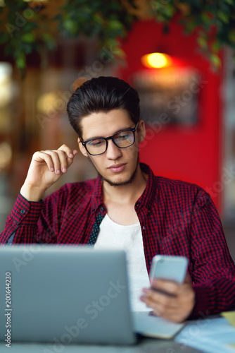 Smiling young hipster man in glasses with smartphone and laptop on the table. Programmer, web developer, designer working at office comparing mobile and desktop website versions. 
