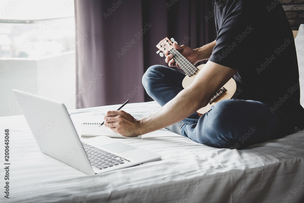 Fototapeta premium Young happiness man on bedroom in enjoying playing the music and compose a song with guitar or ukulele in holiday, relaxation and recreation concept