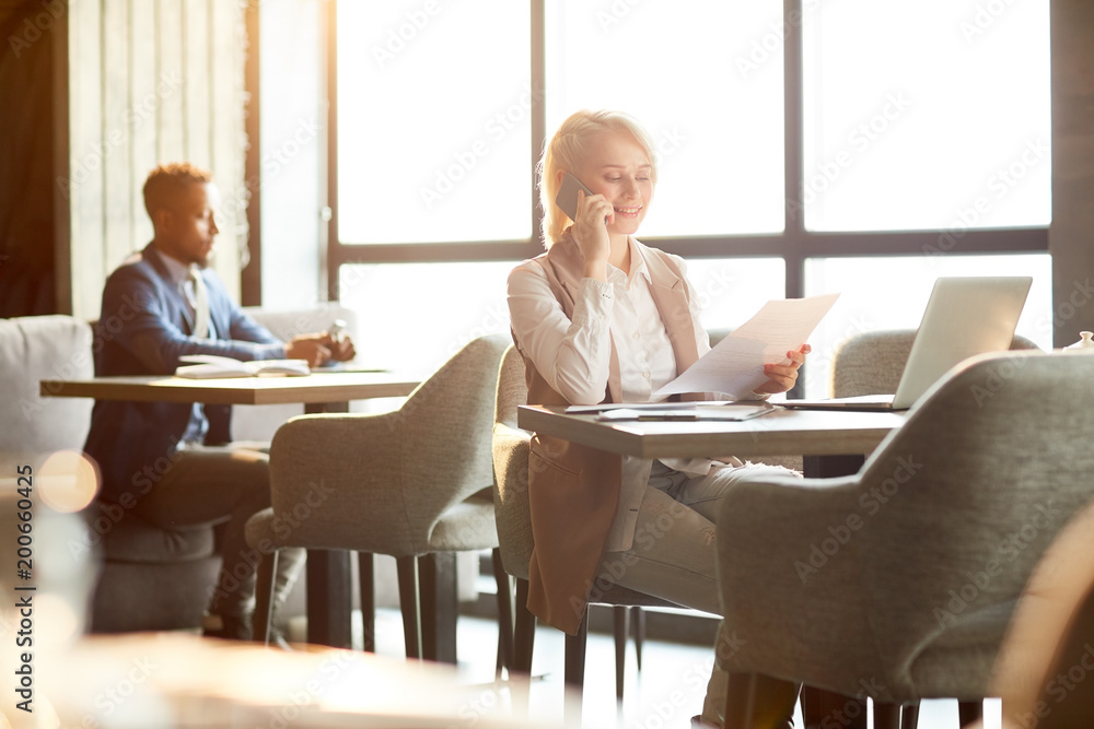 Young agent in smart casual sitting by table in cafe, consulting client by smartphone and looking through text of contract Young agent in smart casual sitting by table in cafe, consulting client by smartphone and looking through text of contract