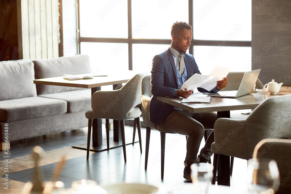 Young elegant business agent sitting by table in cafe and reading financial papers after lunch break Young elegant business agent sitting by table in cafe and reading financial papers after lunch break