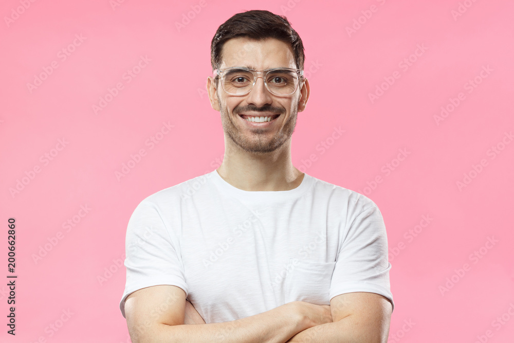 Portrait of smiling handsome man in white t-shirt and transparent eyeglasses standing with crossed arms isolated on pink background Portrait of smiling handsome man in white t-shirt and transparent eyeglasses standing with crossed arms isolated on pink background