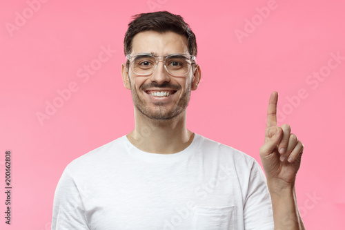 Attractive young man wearing transparent eyeglasses and white t-shirt pointing up with his finger isolated on pink background