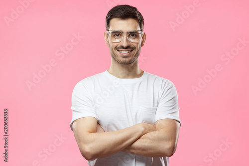 Portrait of smiling handsome man wearing transparent glasses and white tshirt standing with crossed arms isolated on pink background