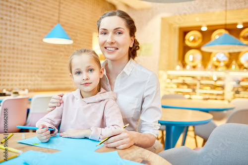 Young woman and little girl with crayons looking at camera while sitting by table in cafe at leisure