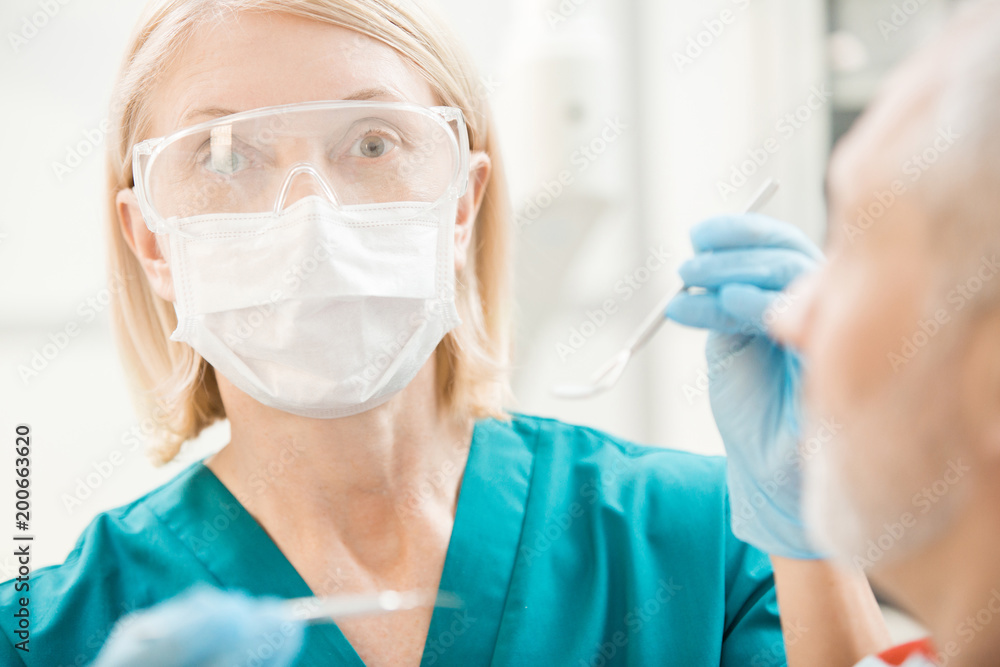 Contemporary dentist in protective mask and eyeglasses going to check up oral cavity of her patient