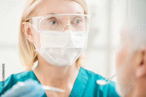 Experienced dentist in protective eyeglasses and mask looking at her patient before oral check-up
