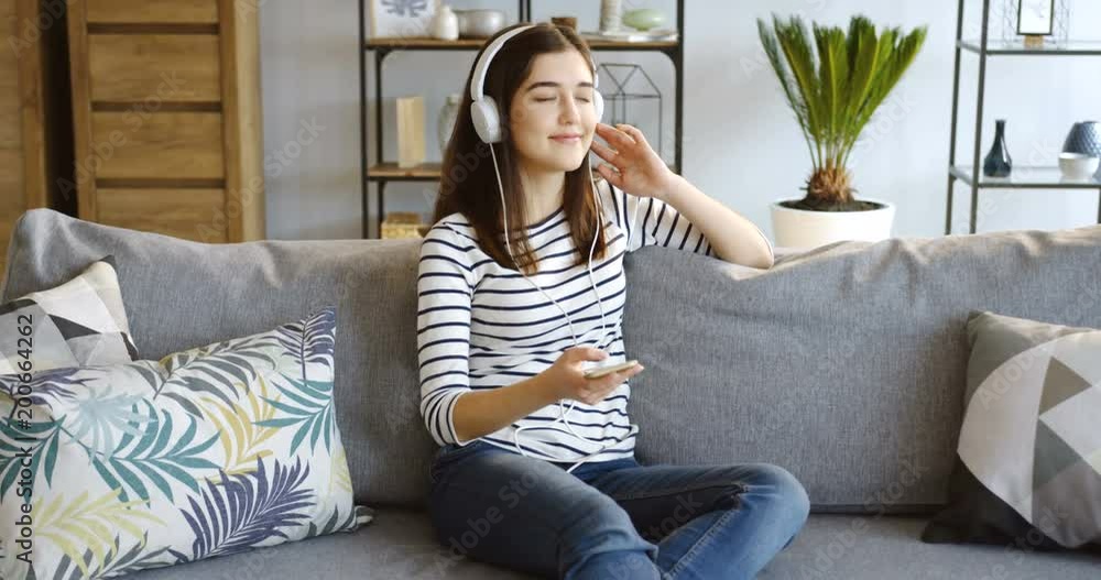 Young charming brunette woman in the striped blouse sitting on the gray couch with pillows, listening to the music on the smartphone via big white headphones and enjoying it in the cozy room at home Young charming brunette woman in the striped blouse sitting on the gray couch with pillows, listening to the music on the smartphone via big white headphones and enjoying it in the cozy room at home