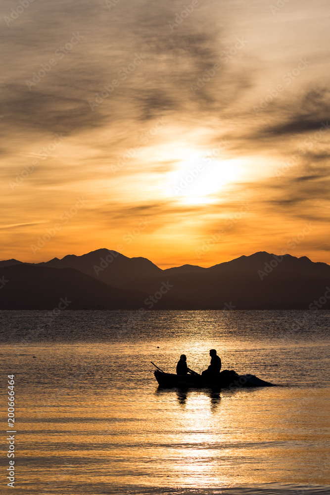Silhouettes of two men rowing in a small fishing boat at a colorful golden cloudy sunrise in Corfu, Greece. Vertical image