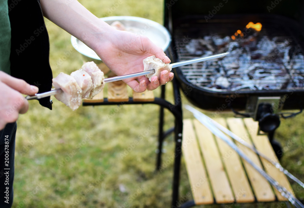 Young teen boy hand put on meat on metal skewer for grilling on flaming grill background in grass on yard outdoors. Concept of summer grilling, picnic, barbecue and party.