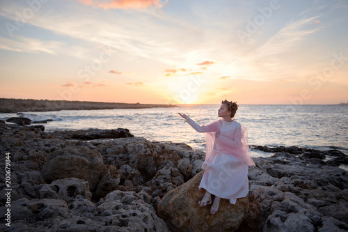 girl crowned princess meets the dawn of the sun sitting on a stone on the ocean sea shore