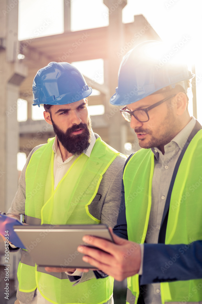 Two young male engineers or business partners at construction site, discussing and using a tablet. Architecture and teamwork concept.