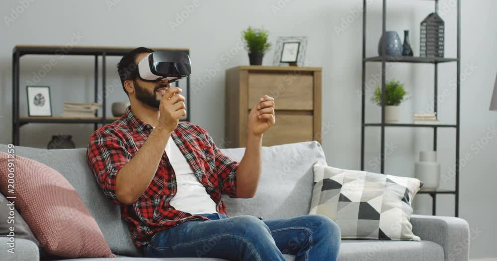 Middle Eastern man in the red plaid shirt sitting on the grey couch in the VR glasses and having VR headset in the living room. Indoors Middle Eastern man in the red plaid shirt sitting on the grey couch in the VR glasses and having VR headset in the living room. Indoors