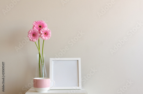 Tall pink gerberas in glass vase with pink mug and blank square frame on small white table against neutral wall background
