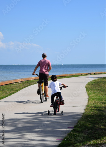 Small boy on bike with training wheels, with father at water front park.