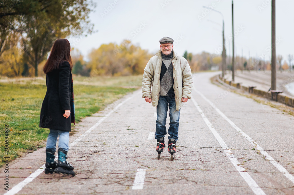 Obraz premium Happy old man goes on rollers with his daughter on the road in autumn park. Happy and active pensioner.