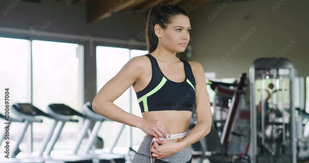 A woman in the gym after various workouts and dieting takes the measuring tape to measure the waistline and see if she has lost weight. Concept of: diet, weight, fitness, sport A woman in the gym after various workouts and dieting takes the measuring tape to measure the waistline and see if she has lost weight. Concept of: diet, weight, fitness, sport
