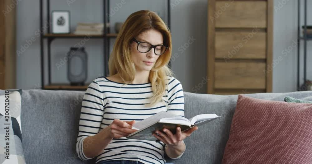 Young blonde pretty woman flipping pages and reading a book on the gray couch in the living room. Indoor Young blonde pretty woman flipping pages and reading a book on the gray couch in the living room. Indoor
