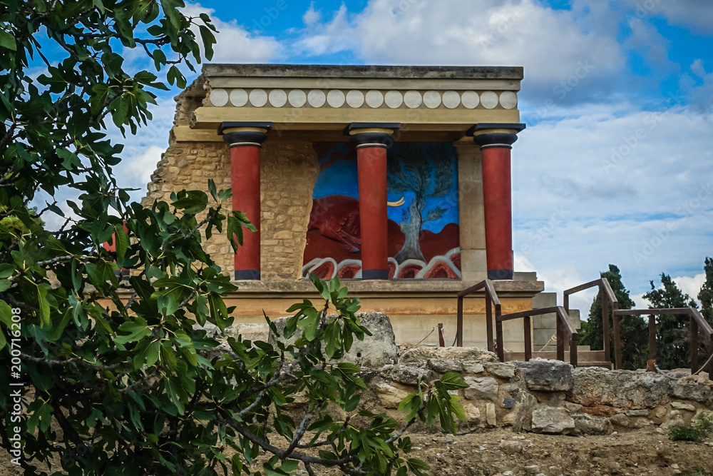 old, historic buildings fragment with red columns and bright wall design on the wall; On the left, in the forefront there are wooden branches with green, robotic leaves, on the back is a blue sky back old, historic buildings fragment with red columns and bright wall design on the wall; On the left, in the forefront there are wooden branches with green, robotic leaves, on the back is a blue sky back