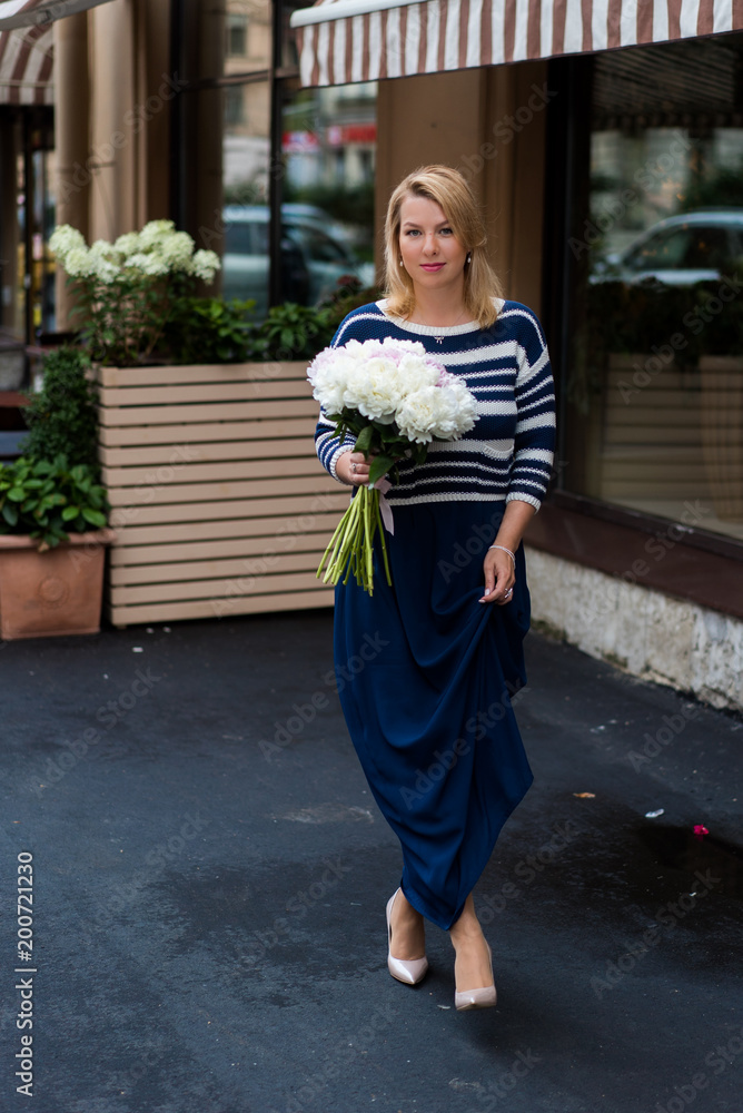 Fototapeta premium Young blonde woman in blue dress with peonies bouquet outdoors at city street
