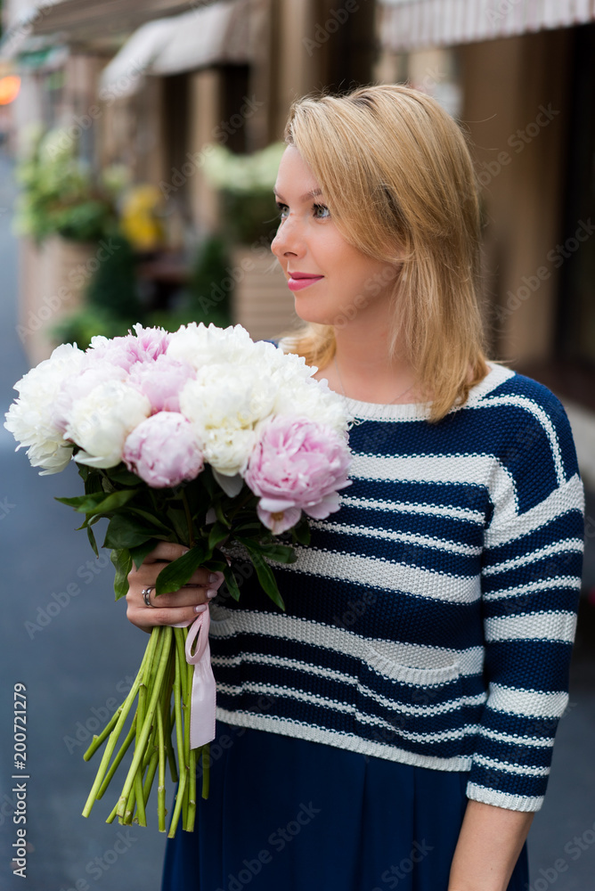 Young blonde woman in blue dress with peonies bouquet outdoors at city street Young blonde woman in blue dress with peonies bouquet outdoors at city street