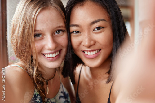 Photo of pleased attractive mixed race women with happy expressions, have broad smiles, stretch hands and make selfie, have interracial relationship. People, friendship and relationship concept