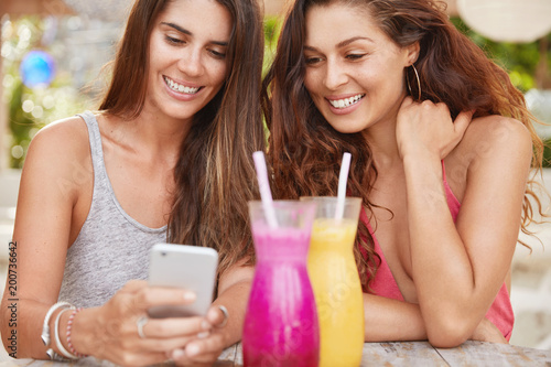 Cropped shot of pleased attractive women have fun together, look with intriguing expression at smart phone as check email box and expect to recieve messagge, recreat together at outside cafeteria