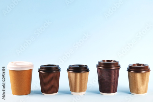 Row of brown carton double walled paper cups, different sizes & colors, take out, coffee to go. Eco-friendly cardboard mugs for hot beverages w/ cap, sky blue gradient background, close up, copy space