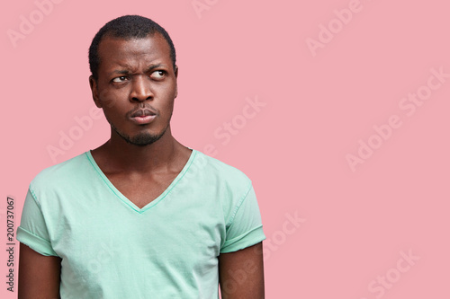 Horizontal shot of thoughtful young attractive African American male with dark skin, dressed in casual t shirt, poses against pink background with copy space for your advertisment or promotional text