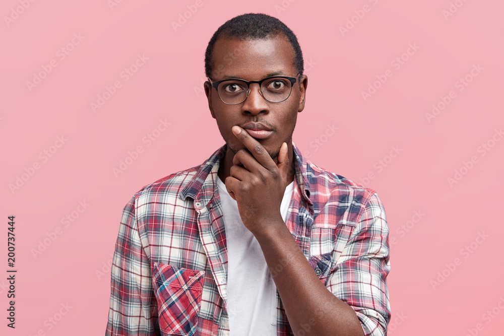 Headshot of serious dark skinned male looks scrupulously at camera, being confident, wears spectacles and checkered shirt, isolated over pink background. Handsome man poses indoor. Facial expressions Headshot of serious dark skinned male looks scrupulously at camera, being confident, wears spectacles and checkered shirt, isolated over pink background. Handsome man poses indoor. Facial expressions