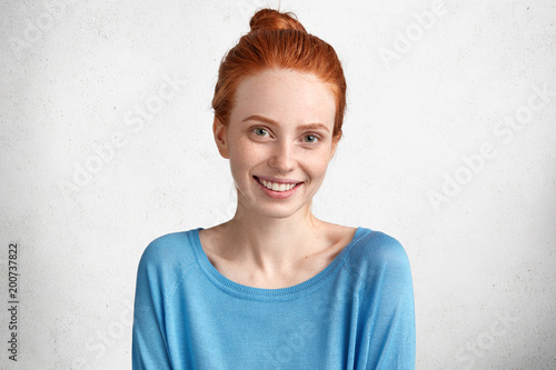 Glad smiling female with freckled skin and positive smile, dressed in blue casual sweater, being in high spirit after date with boyfriend, isolated over white background. Beautiful red haired woman