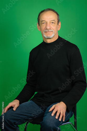 Handsome Gray-haired Mature Man with Well-trimmed Beard in Black Sweater and Dark Jeans Sitting on the Chair on a Green Background in Studio